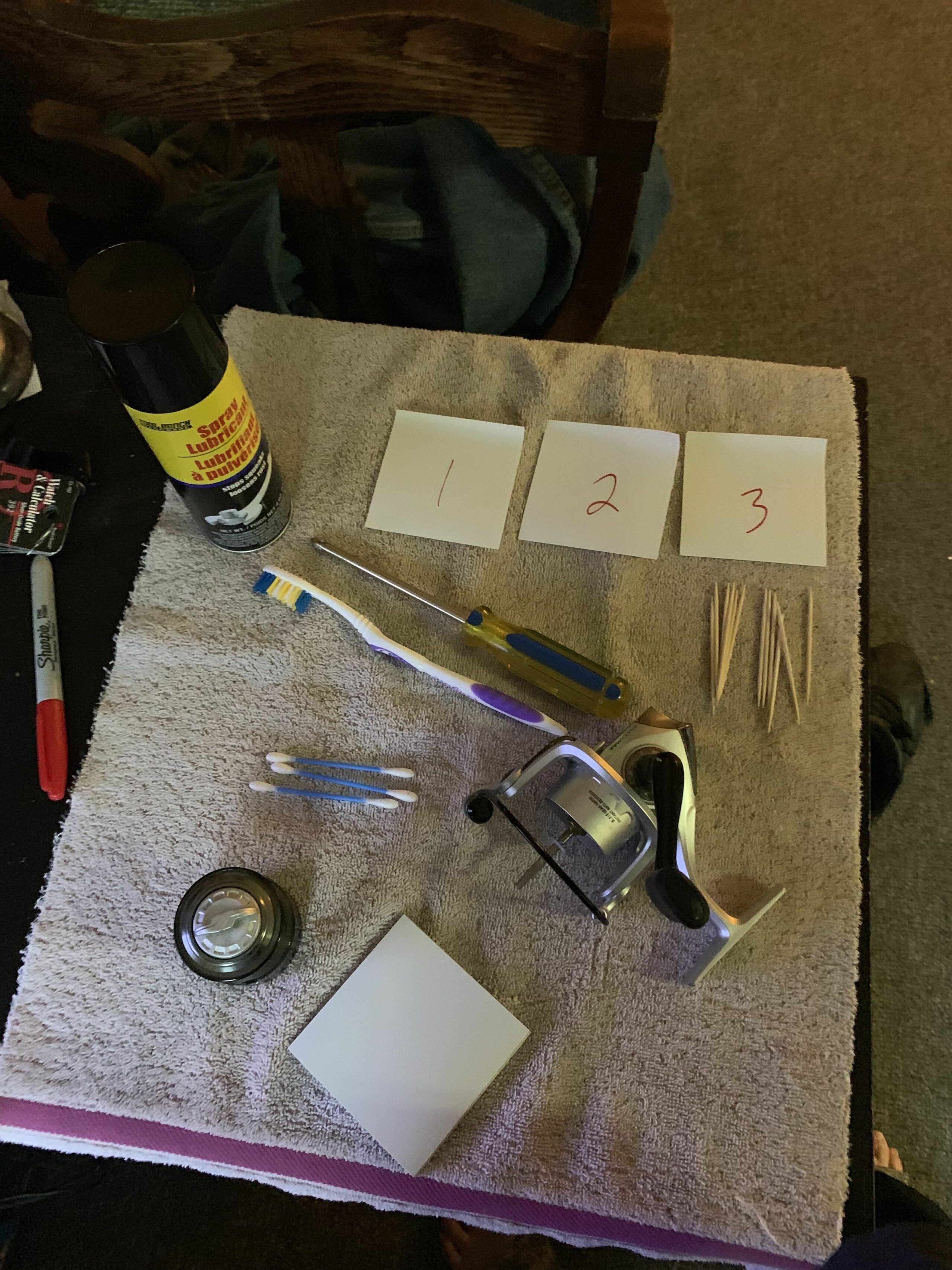 Items use for cleaning a spinning reel. Spinning fishing reel with spool detached. Toothbrush, phillips head screwdriver, toothpicks q-tips, 3 sticky notes 1,2,3 written on them, lubricant cleaner, towel.