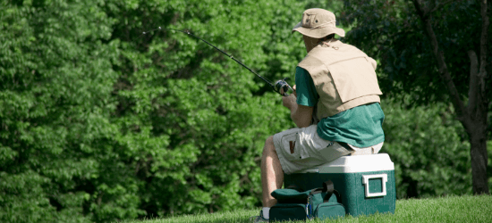 Man sitting on a cooler with a fishing pole and bag next to him. Trees and grass in the picture.