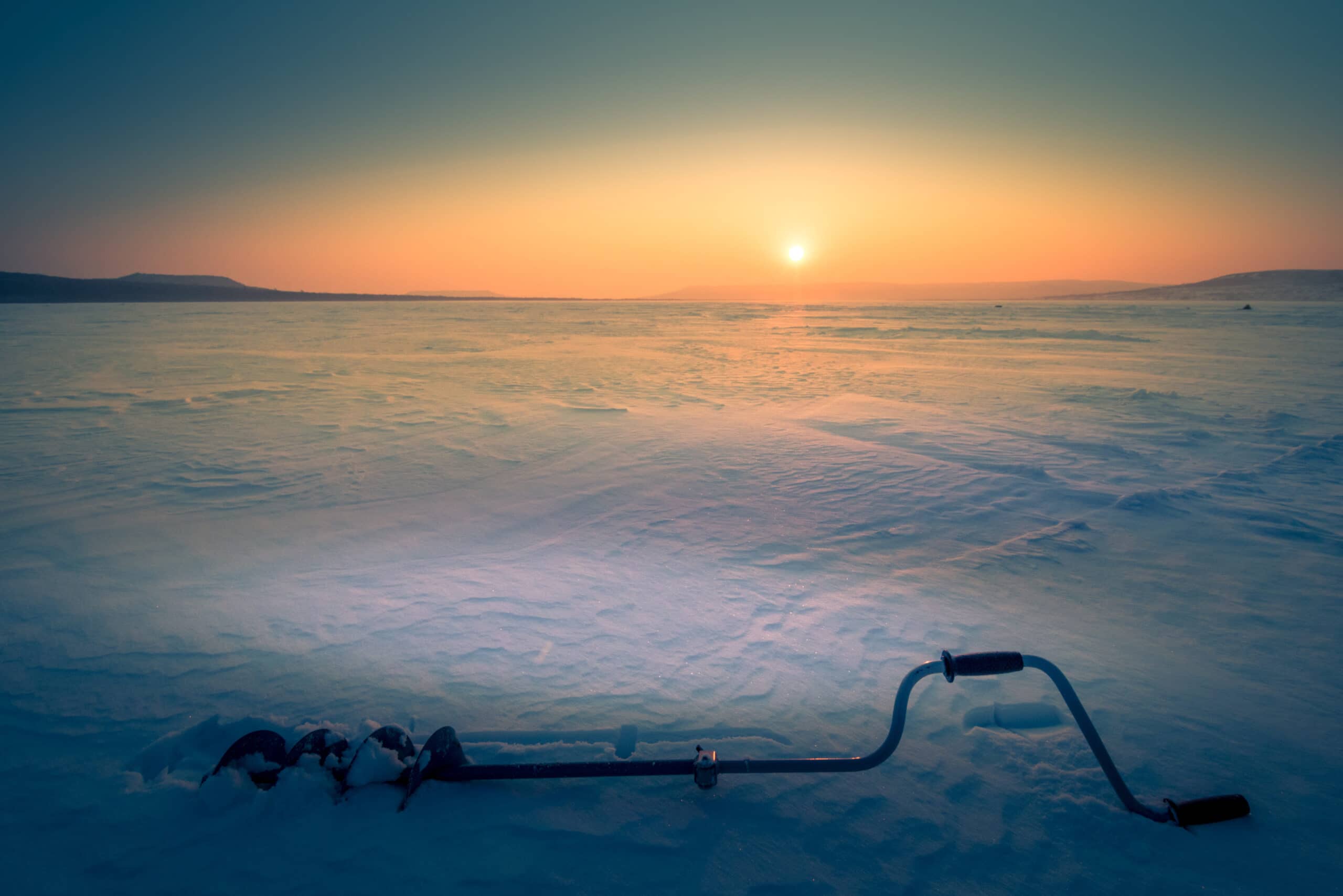 Frozen lake with sun in background and an ice auger in the foreground.